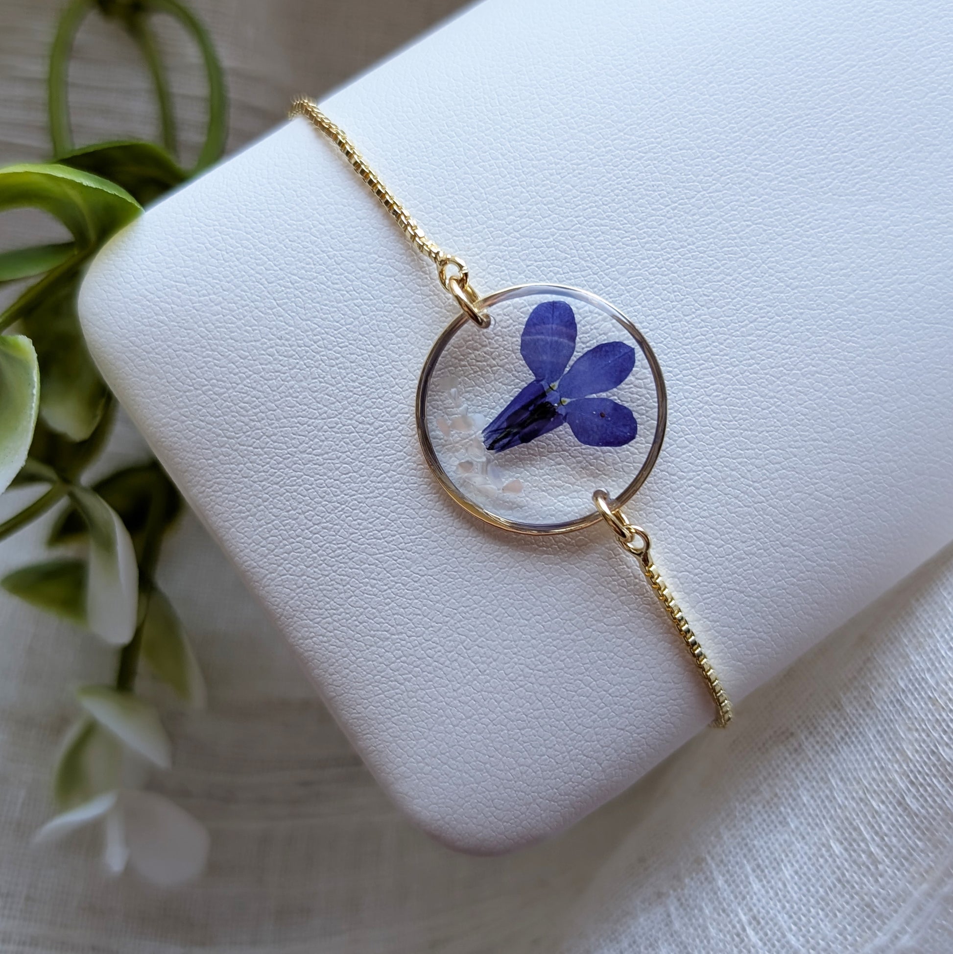 Gold plated blue lobelia bracelet shown on a jewellery holder, crafted with garden grown flowers, resin and seashell flakes