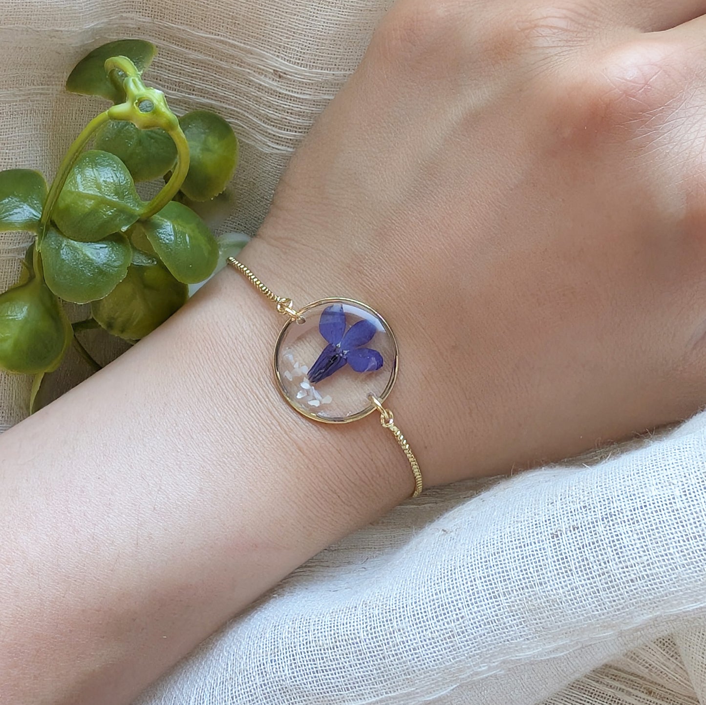 Model with fair skin wearing a blue lobelia 20mm flat bracelet, delicate gold plated real flower jewellery for everyday wear