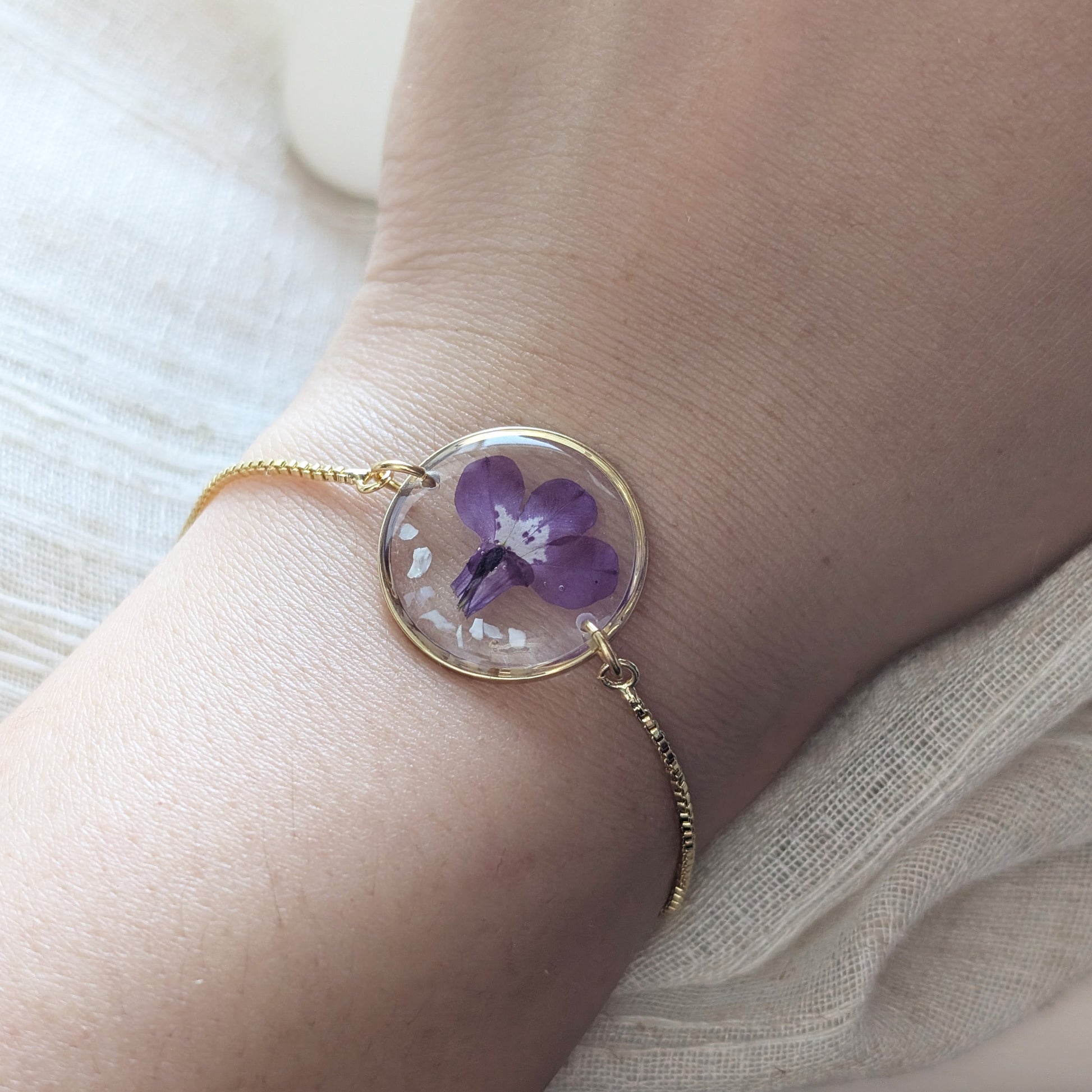 Model with fair skin wearing a purple lobelia 20mm flat bracelet, delicate gold plated real flower jewellery for everyday wear